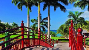 Un puente de madera con barandillas rojas que lleva a una playa con palmeras y un cielo azul despejado.