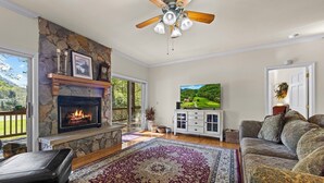 A living room with a stone fireplace, a ceiling fan, a television, and a patterned rug. - Condo w/ Creek, Golf & Mtn Views, Only 5 Steps to get in and Washer & Dryer! (Whittier)