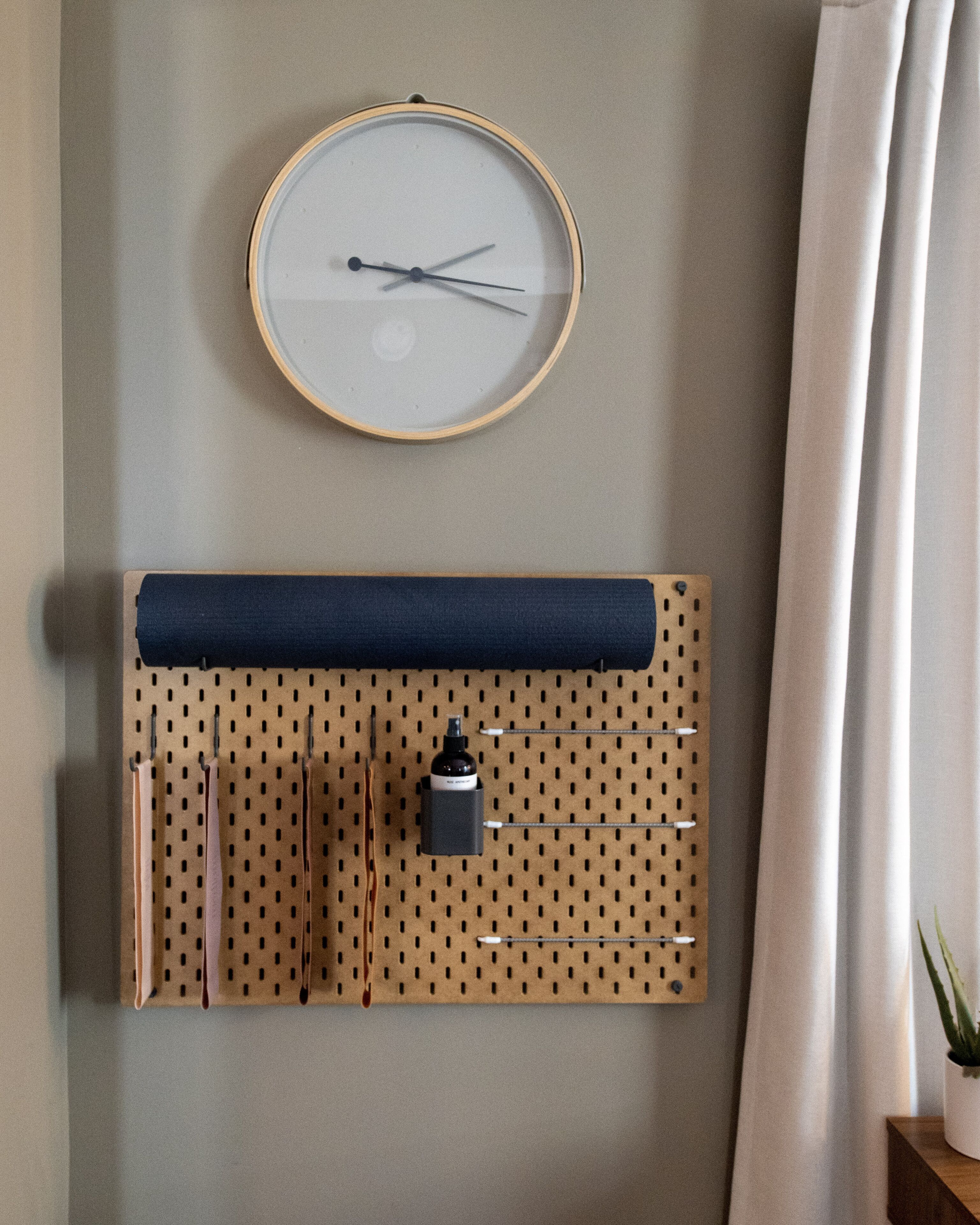 Eine an der Wand befestigte Uhr mit Holzrahmen, darunter eine Siebdrucktafel und eine Zimmerpflanze rechts.