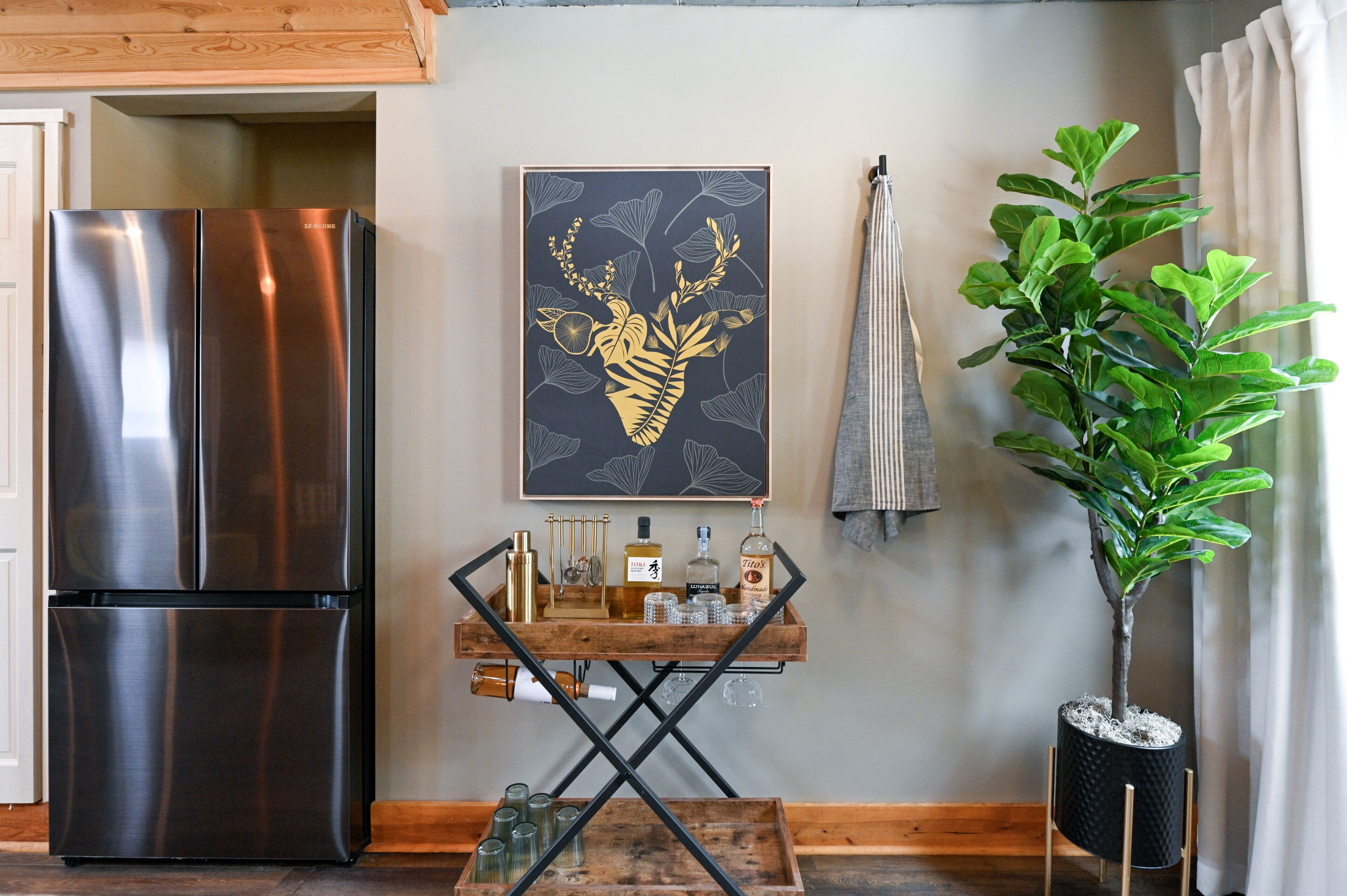 A modern kitchen with a stainless steel refrigerator, a wooden serving cart with bottles and glasses, and a potted plant.