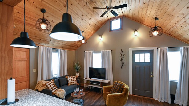 Ein gemütliches Wohnzimmer mit einer Couch, einem Sessel und einem Fernseher, Holzboden und großen Fenstern mit Vorhängen.