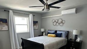 Un dormitorio con una cama, dos mesitas de noche, un ventilador de techo, una ventana con cortinas y un aire acondicionado instalado en la pared.