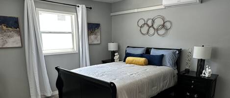 A bedroom with a bed, two bedside tables, a ceiling fan, a window with curtains, and a wall-mounted air conditioner.