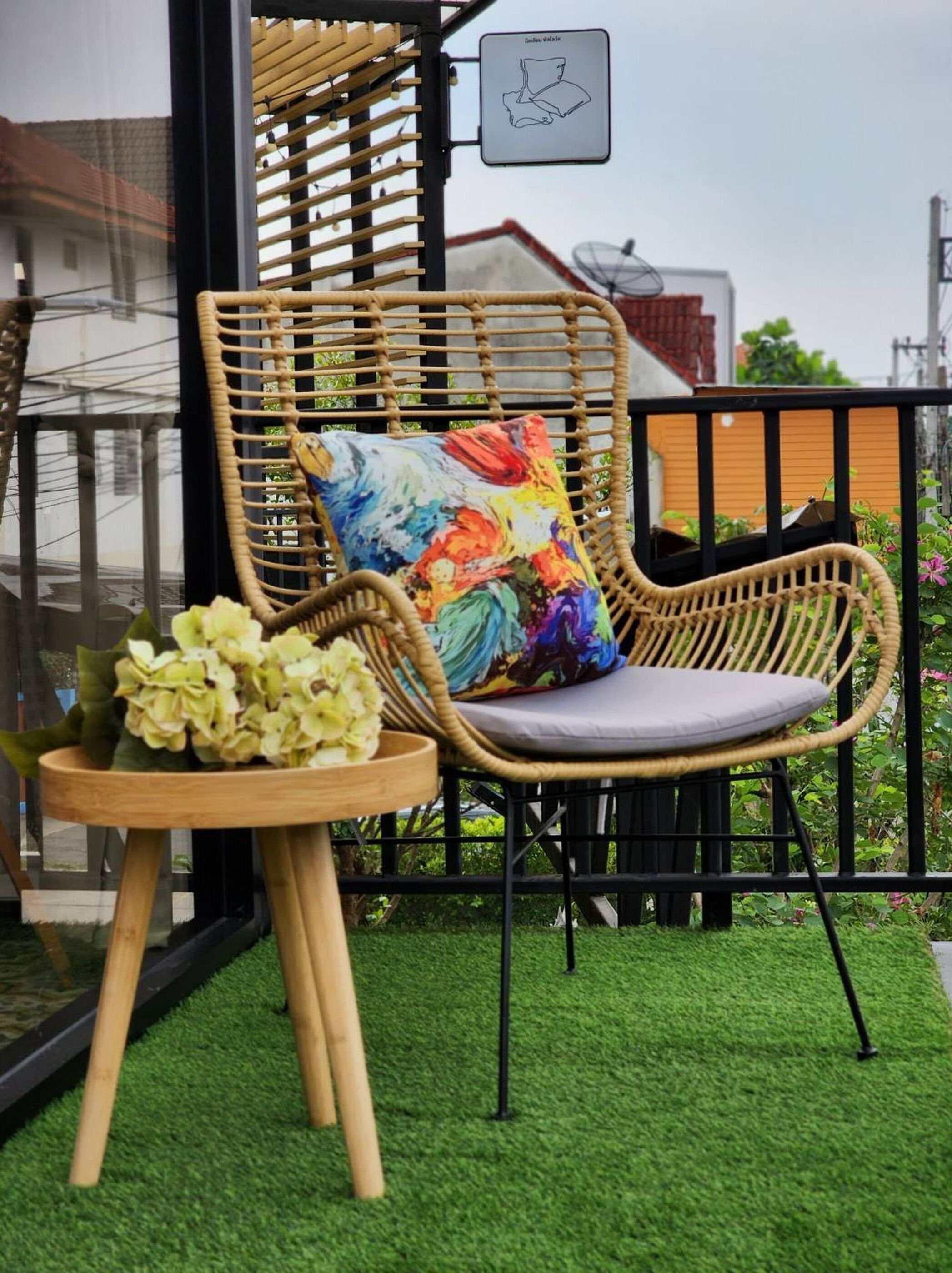 A wicker chair with a colorful cushion, a small wooden table with flowers, and a balcony railing.