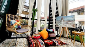 A room with a large window, a wicker chair with a colorful cushion, a small table with a painting, and patterned floor tiles. - Capital O 75422 Million Pillows  (Chiang Mai)