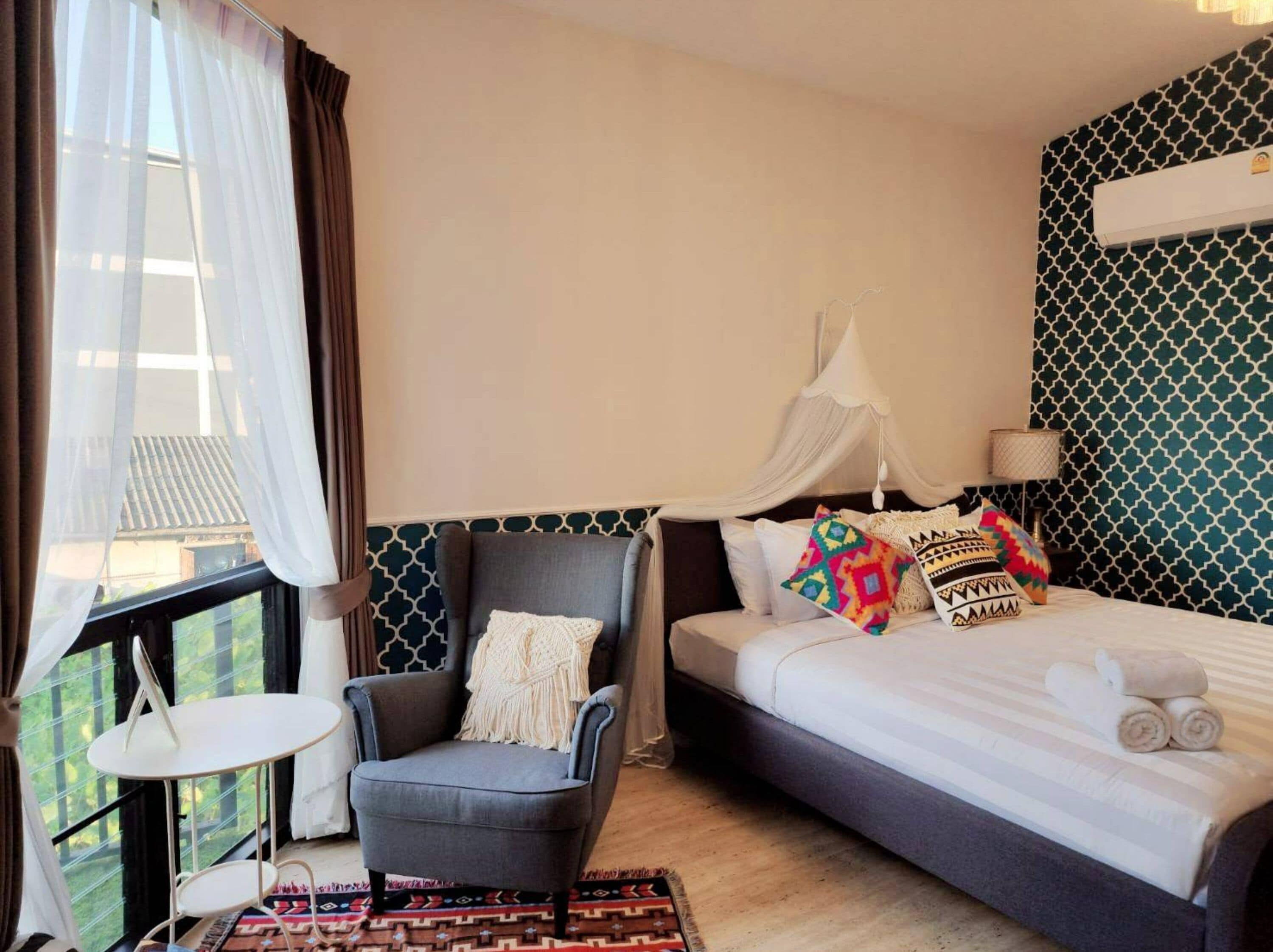 A hotel room with a bed, a chair, a small table, and a window with curtains.