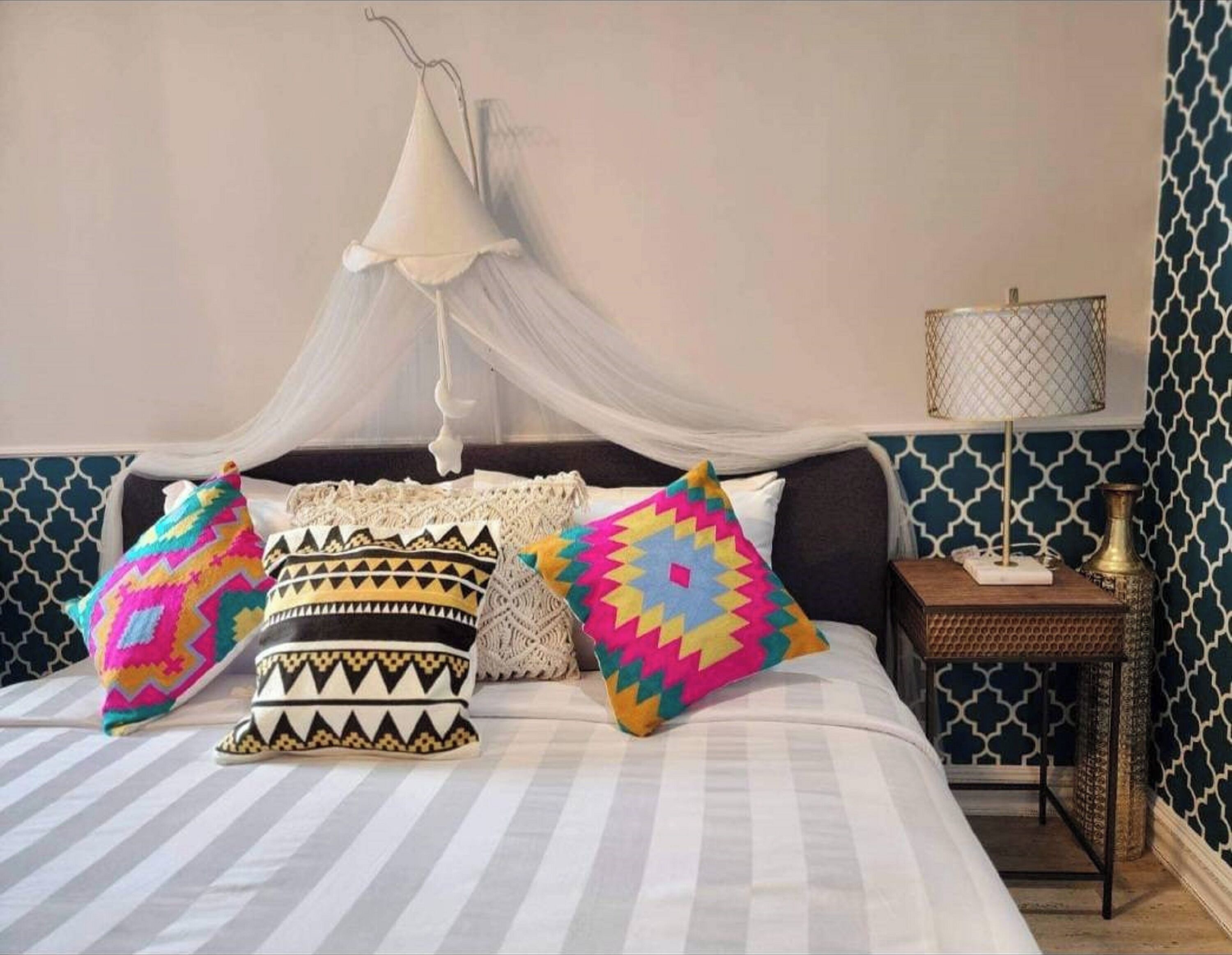 Un lit avec des coussins à motifs, une table de chevet avec une lampe et un baldaquin au-dessus.