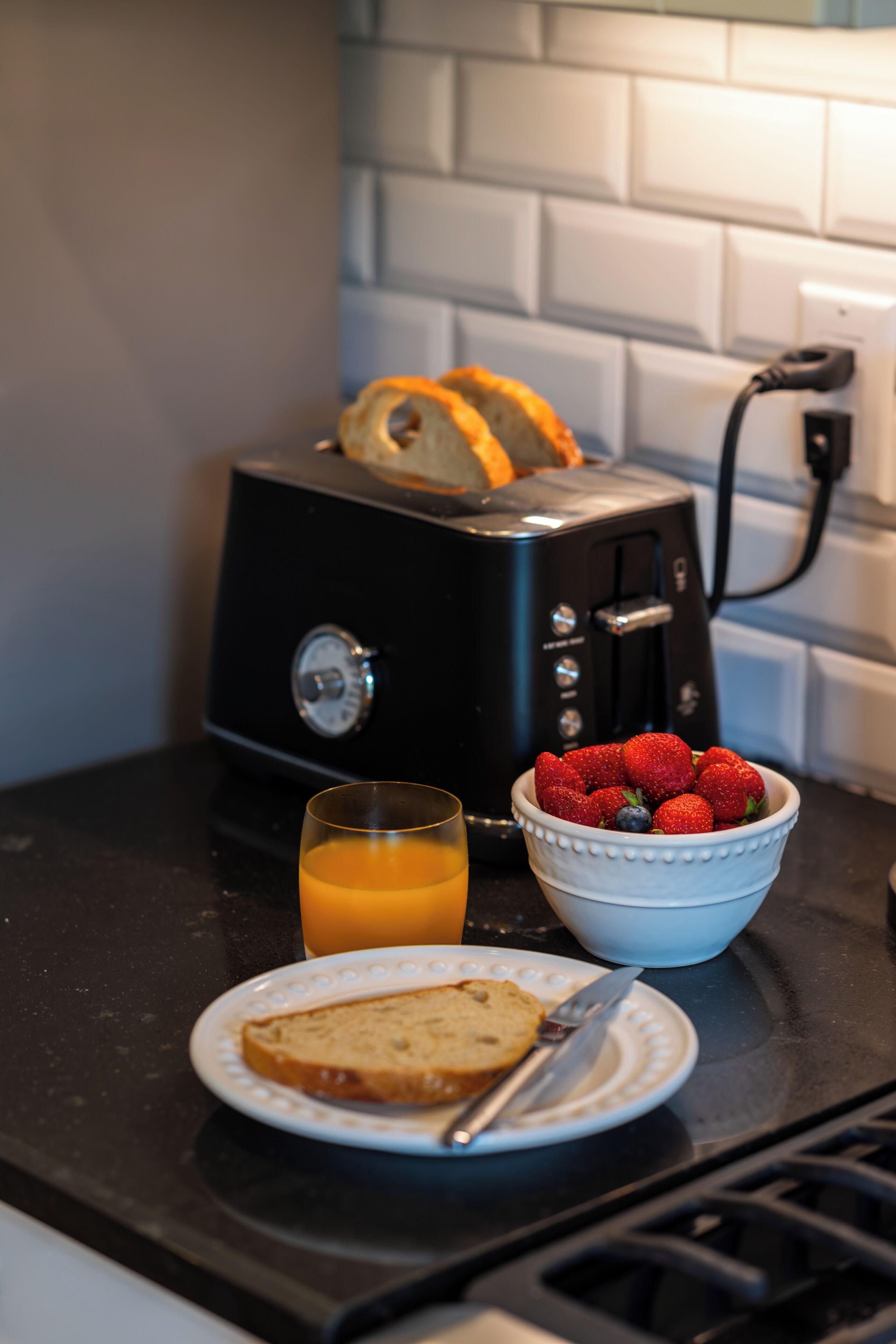 A kitchen counter with a toaster, a bowl of strawberries, a glass of orange juice, and a plate with toast and a knife.