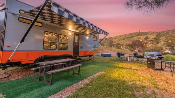 A recreational vehicle with a checkered awning, a picnic table, and a barbecue grill set up in a grassy area with a hilly backdrop. - Harley Davidson Ranch Glampling & Kids Play Area (Ramona)