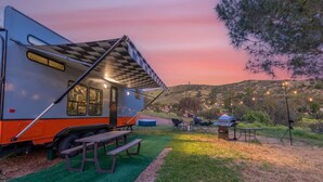 A recreational vehicle with a picnic table and barbecue grill set up in a grassy area with a scenic view of hills and a sunset. - Harley Davidson Ranch Glampling & Kids Play Area (Ramona)