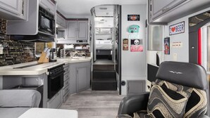 A compact RV kitchen with a microwave, stove, and sink. There is a bed in the background and a sign on the wall that says "I love beer." - Harley Davidson Ranch Glampling & Kids Play Area (Ramona)