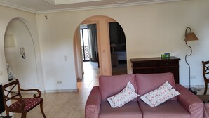 Sala de estar con un sofá rosa, sillas de madera, televisor y lámpara. Hay dos arcos que dan a otra habitación.