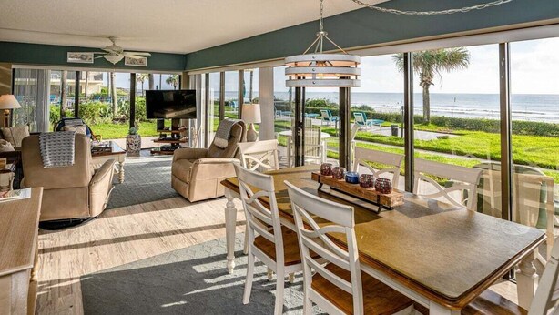 A spacious living area with a large window overlooking the ocean, a television, and comfortable seating. - Coastal Haven at Ocean House 120 (St. Augustine)
