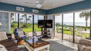 A spacious living room with a large TV, comfortable seating, and a view of the ocean through large sliding glass doors. - Coastal Haven at Ocean House 120 (St. Augustine)