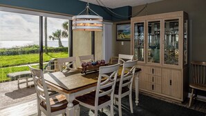 A dining area with a wooden table and white chairs, a glass cabinet with glassware, and a view of the ocean through large windows. - Coastal Haven at Ocean House 120 (St. Augustine)