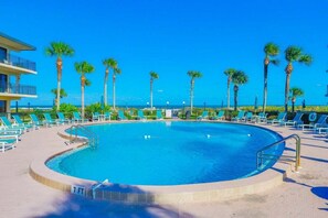 Ein Pool mit einer 2,1-Meter-Tiefenmarkierung, umgeben von Liegestühlen und Palmen, mit einem Gebäude im Hintergrund.