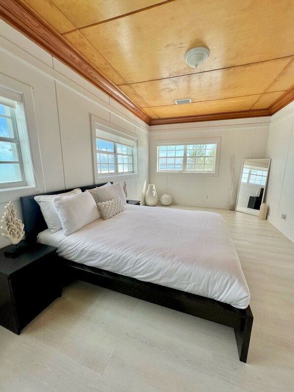 A spacious bedroom with a large bed, wooden ceiling, and multiple windows providing natural light.
