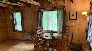 A wooden cabin with a round dining table, six wooden chairs, checkered curtains, a clock, a framed picture, and a table lamp. - Sugarloaf camp - ski/ outdoors rustic retreat (Carrabassett valley)