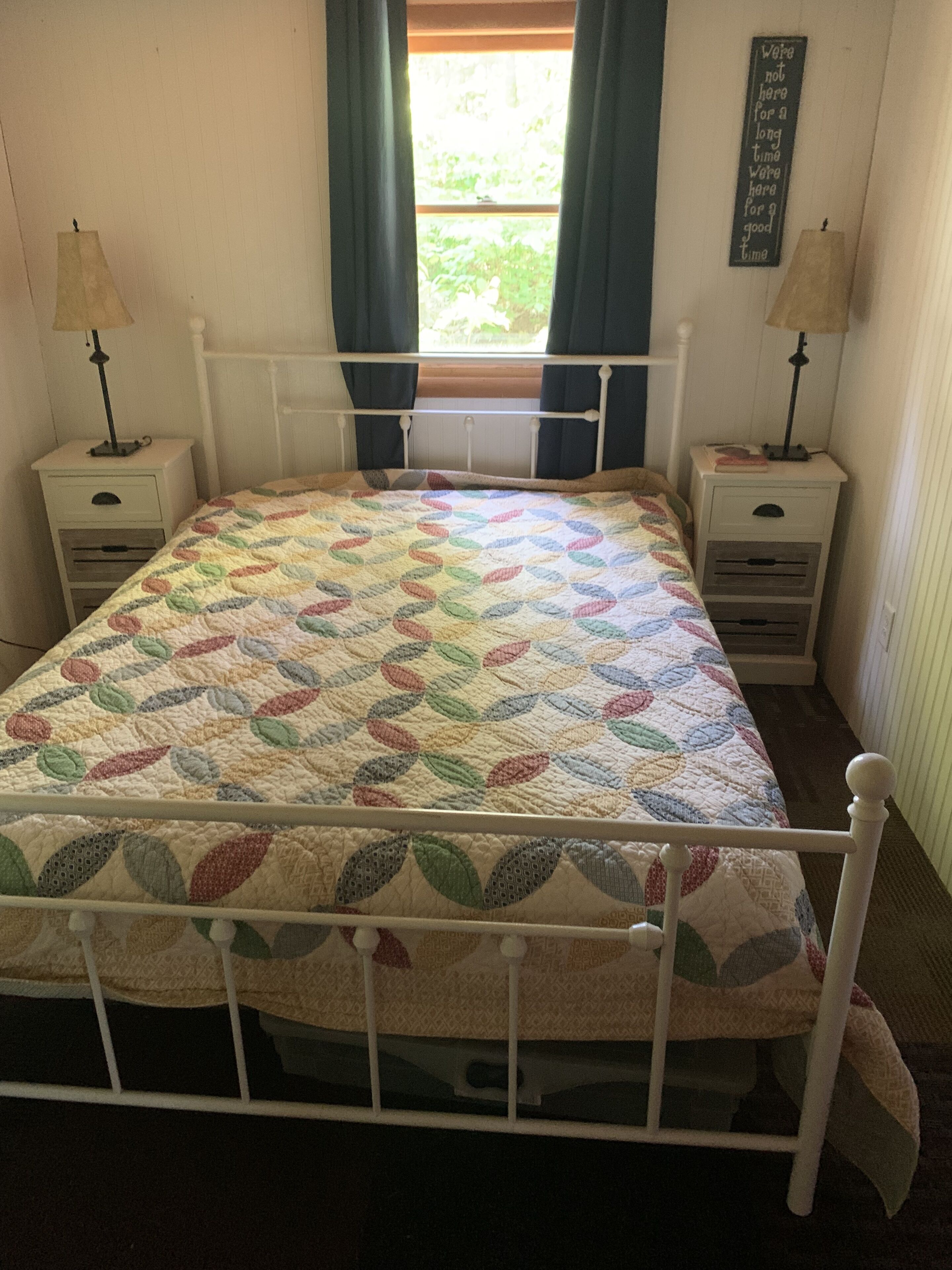 A bed with a colorful leaf-patterned quilt, two bedside tables with lamps, a window with curtains, and a wall-mounted sign.