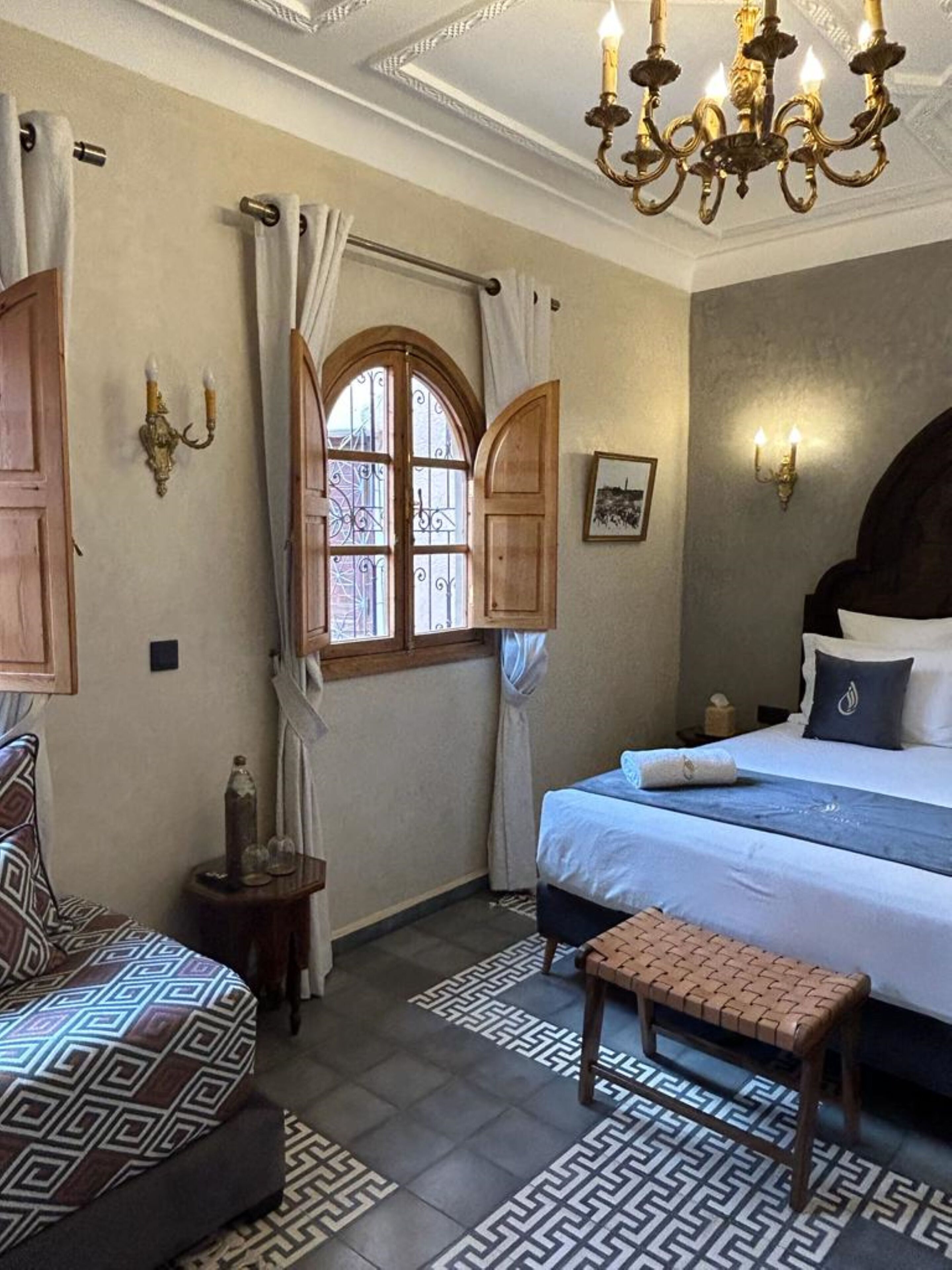A bedroom with a bed, a bench, a window with curtains, and a chandelier.