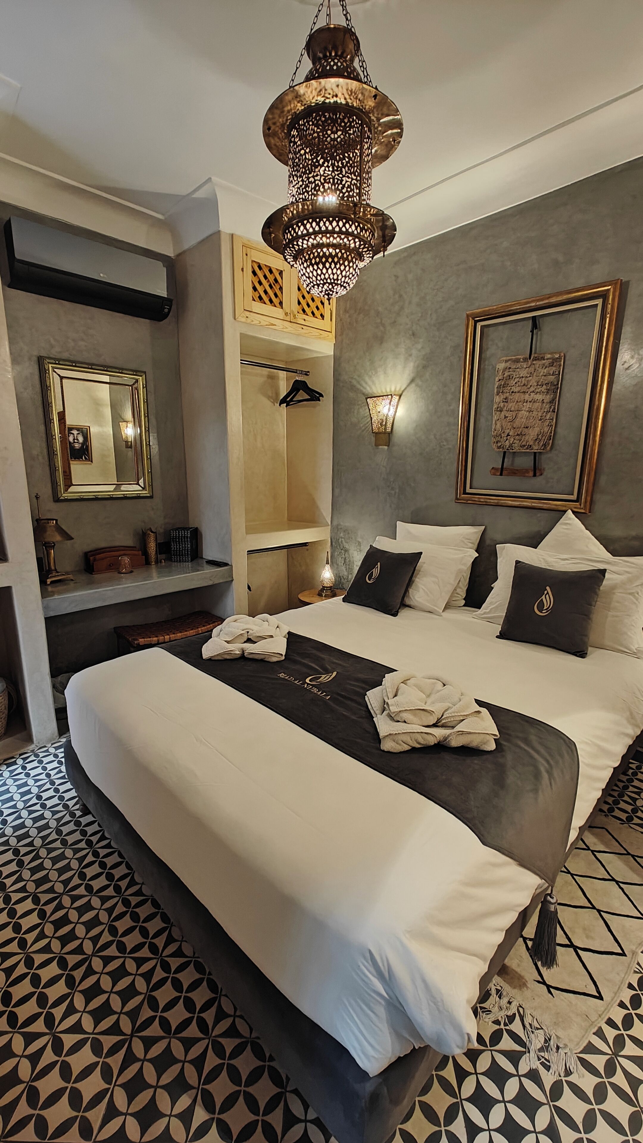 A modern hotel room with a large bed, a hanging light fixture, a wardrobe, and a wall-mounted mirror.