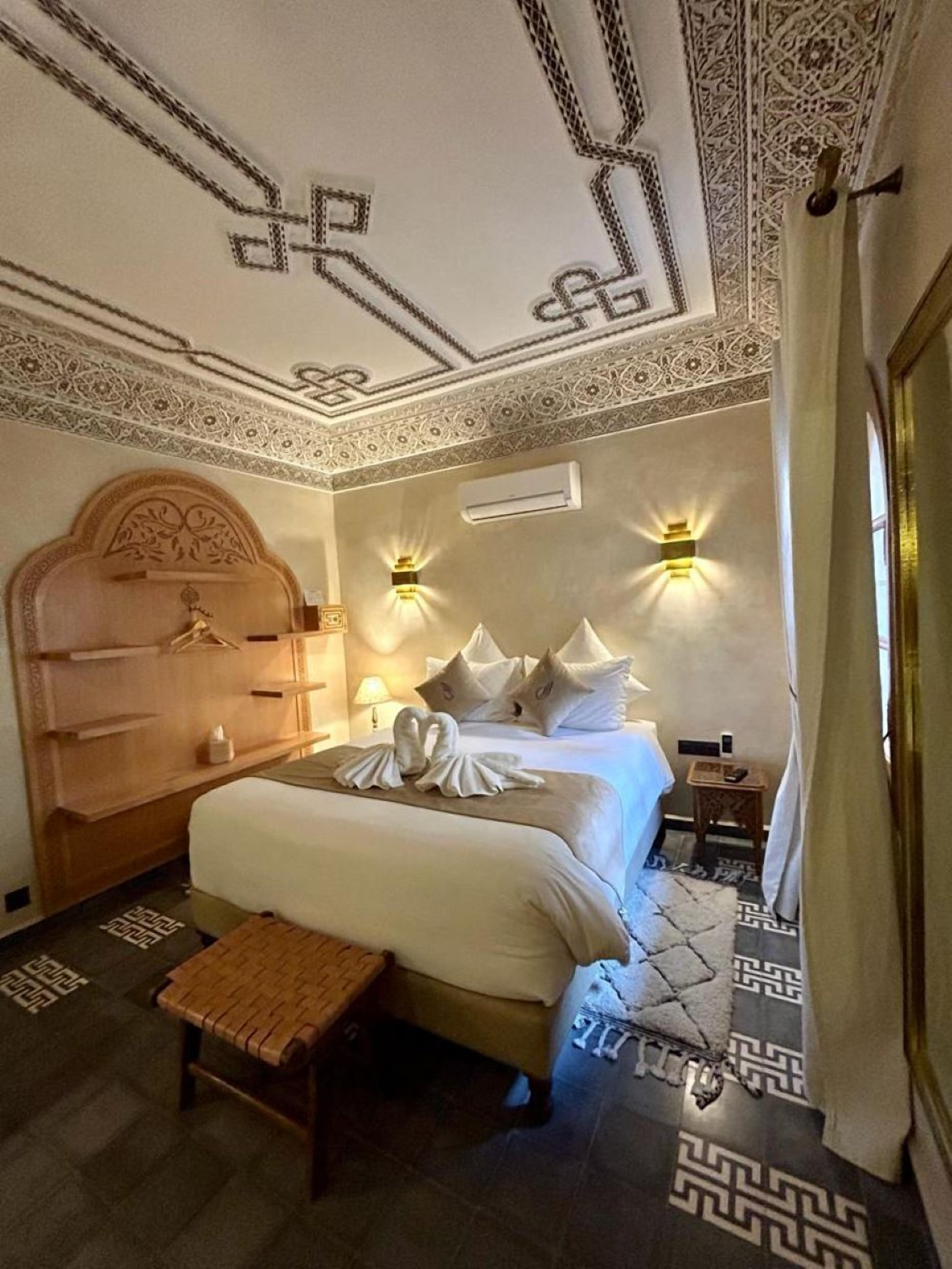 A hotel room with a large bed, decorative ceiling, wall sconces, and a wooden bench.