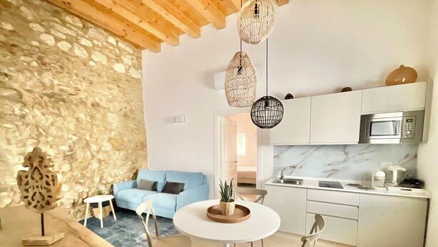 A modern kitchen with white cabinets, a microwave, and a marble backsplash. There is a dining area with a round table and chairs, and a living room with a blue sofa and a stone wall. - RUA 39 fine apartments 3 (Syracuse)