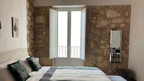 A bedroom with a stone wall, a bed with a striped bedspread, two pillows, a chair, and a mirror on the wall. - RUA 39 fine apartments 3 (Syracuse)