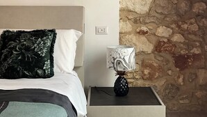 A bedroom with a stone wall, a bed with a floral pillow, a nightstand with a lamp, and a painting on the wall. - RUA 39 fine apartments 5 (Syracuse)