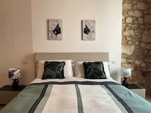 Un dormitorio con cama, dos almohadas, una mesita de noche, una lámpara y una pared de piedra.