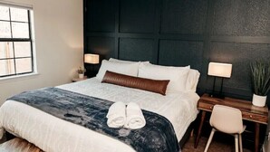 A neatly made bed with a dark headboard, two bedside lamps, a small desk with a chair, and a window with blinds. - 2 minutes to downtown! Hill Country Luxury Duplex TX Wine Trail - Home B (Johnson City)