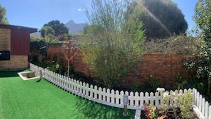 A backyard with artificial green turf, a white picket fence, and a brick wall. - Mispa George - Self Catering Apartment - Sleeps 2 (George)