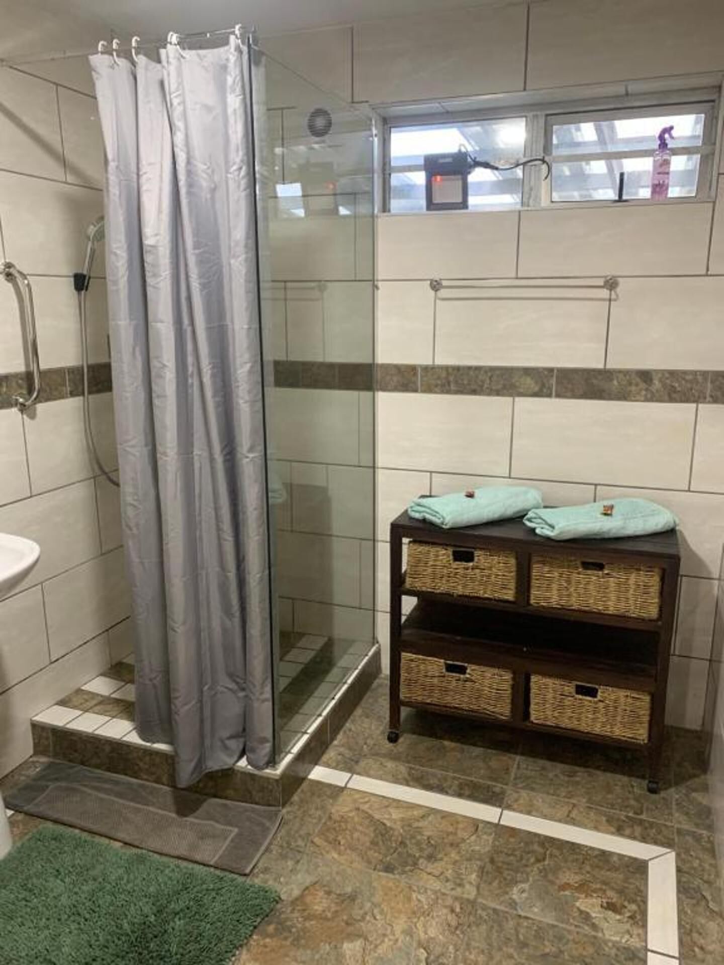 A bathroom with a shower, a sink, a small table with baskets, and a window with a view.