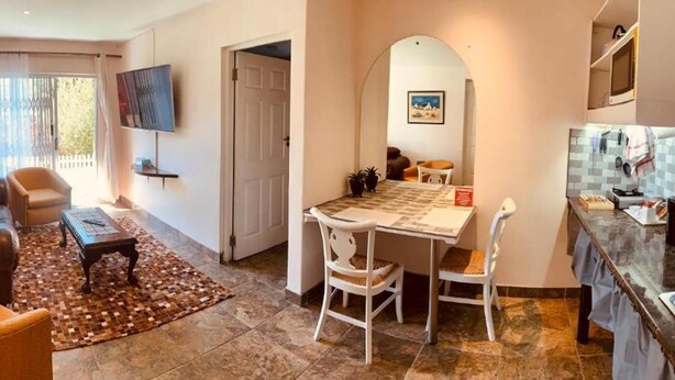 A living room with a sofa, armchair, and coffee table. There is a dining area with a table and chairs. A TV is mounted on the wall. - Mispa George - Self Catering Apartment - Sleeps 2 (George)