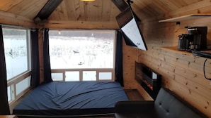 A cozy cabin room with a bed, a fireplace, a coffee maker, and a large window overlooking a snowy landscape.