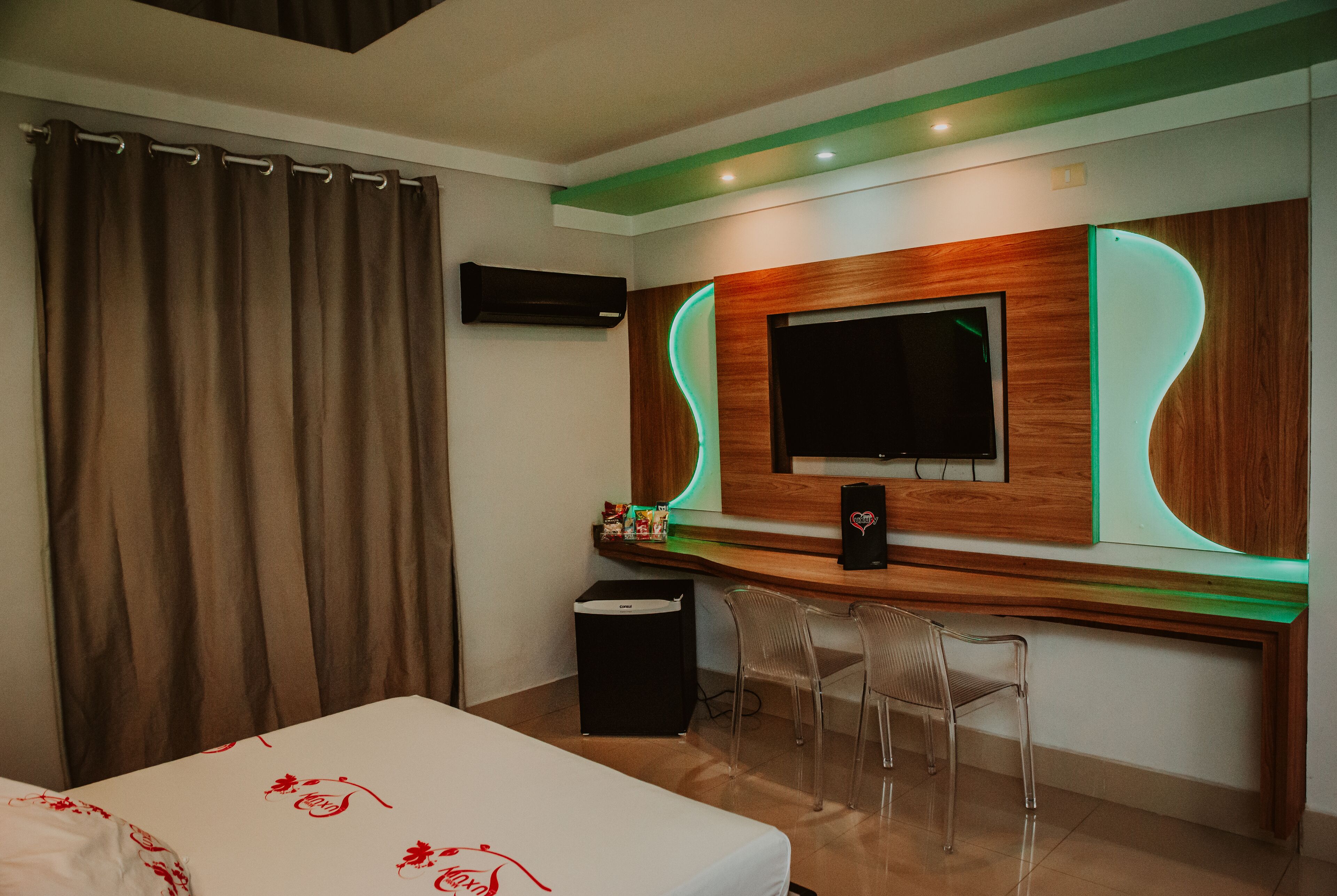 A hotel room with a bed, a TV mounted on a wooden wall, a desk with a computer, and two chairs.