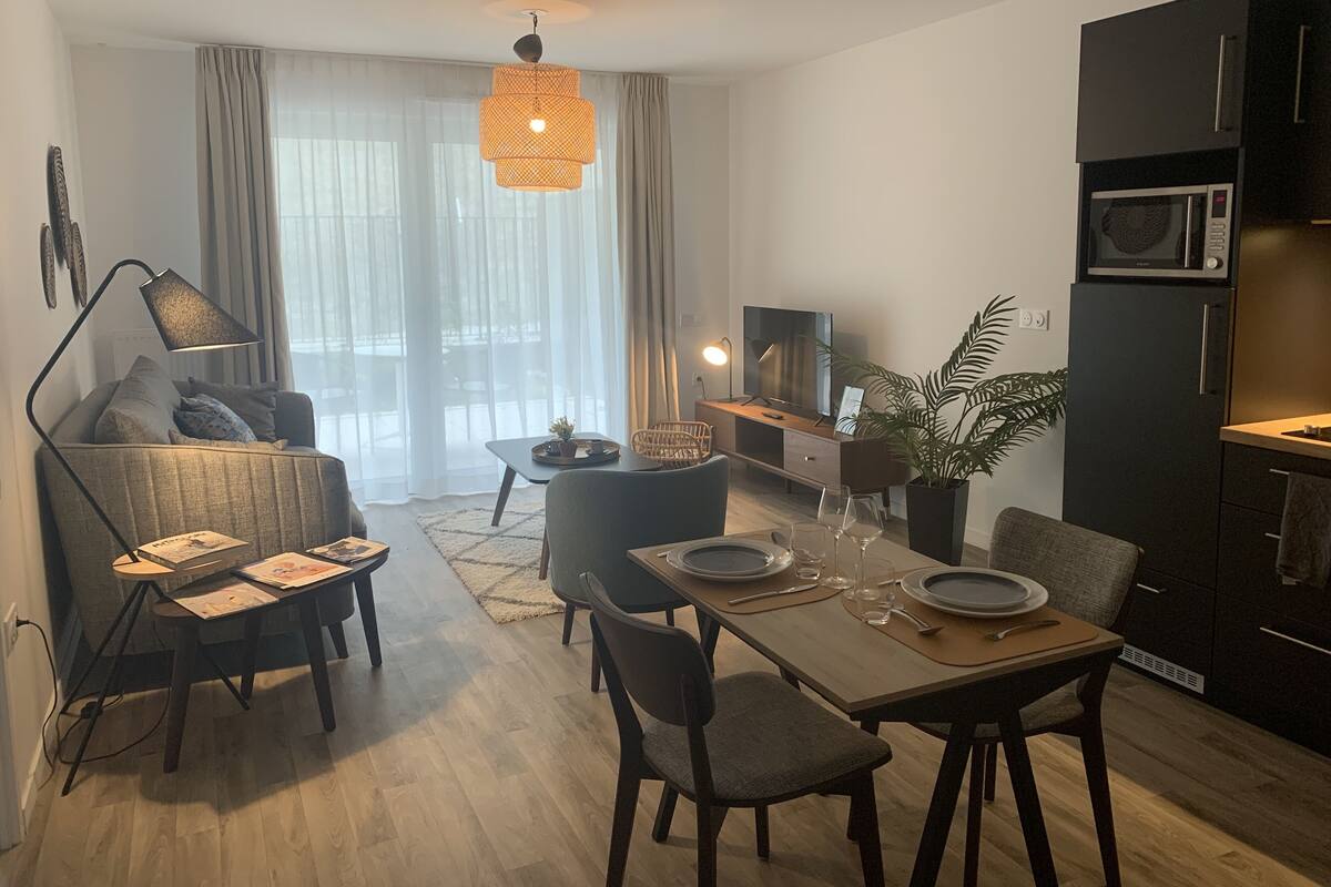 A modern living room with a sofa, dining table, and chairs. There is a television, a plant, and a kitchen area with a microwave.