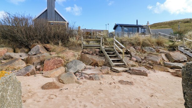 Ein sandiger Strand mit einer Holztreppe, die zu einem Gebäude mit grauem Dach führt.