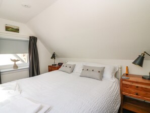 A neatly made bed with white linens, a wooden nightstand with a lamp, and a window with a view of buildings. - Shell Cottage (Peterhead)