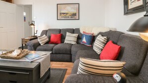 Ein gemütliches Wohnzimmer mit grauem Sofa, gestreiften Kissen, einer roten Decke und einem gerahmten Reiseplakat an der Wand.