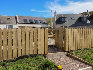 A wooden gate leading to a gravel pathway with a garden bed and a yellow flower. - Shell Cottage (Peterhead)