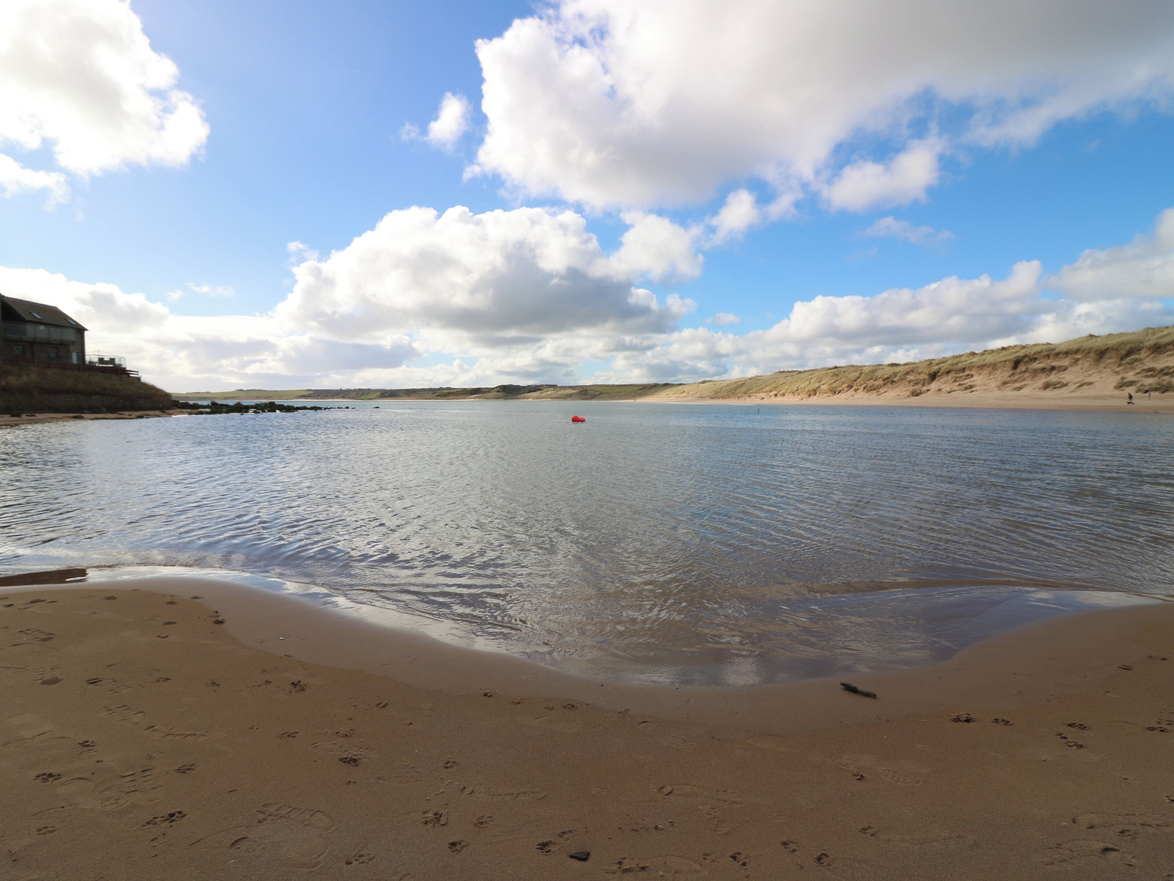 Une plage parsemée d’empreintes, une étendue d’eau calme et un bâtiment visible au loin.