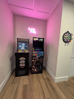Two arcade machines in a room with a neon sign reading 'GAME ZONE' and a dartboard on the wall.