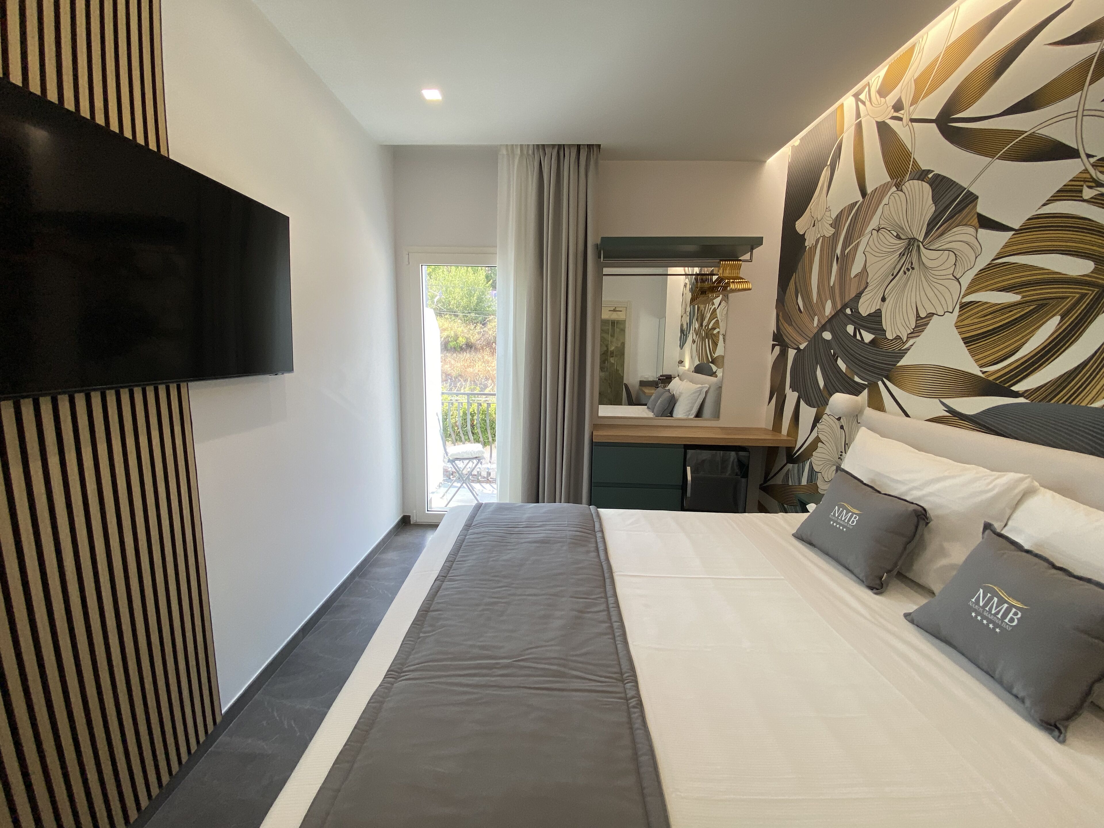 A modern bedroom with a large bed, a flat-screen TV mounted on the wall, a desk with a chair, and a large window with a view of greenery.