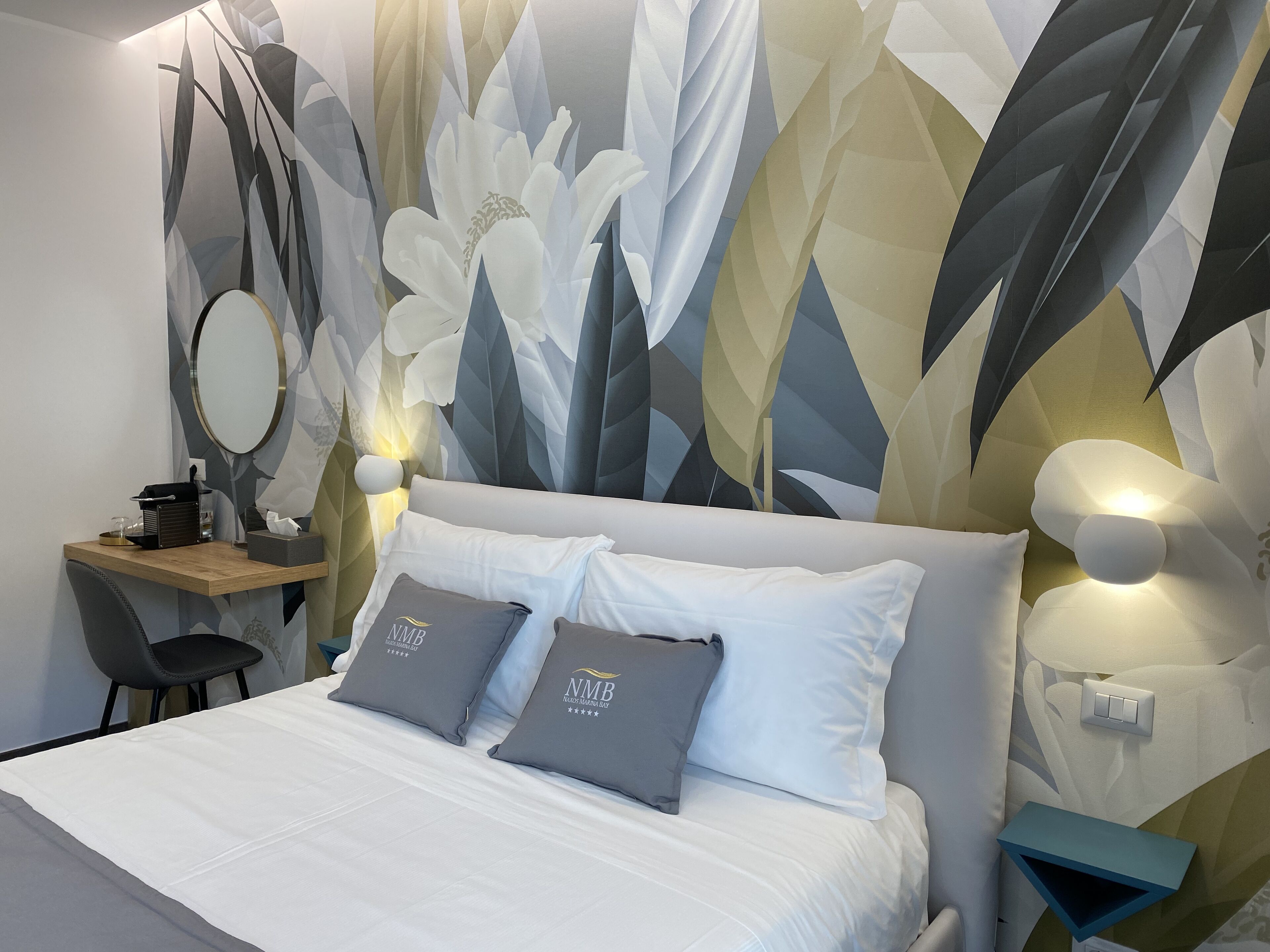 Una cama bien tendida con dos almohadas, una mesita de noche con una lámpara, una silla y una pared con estampado floral.
