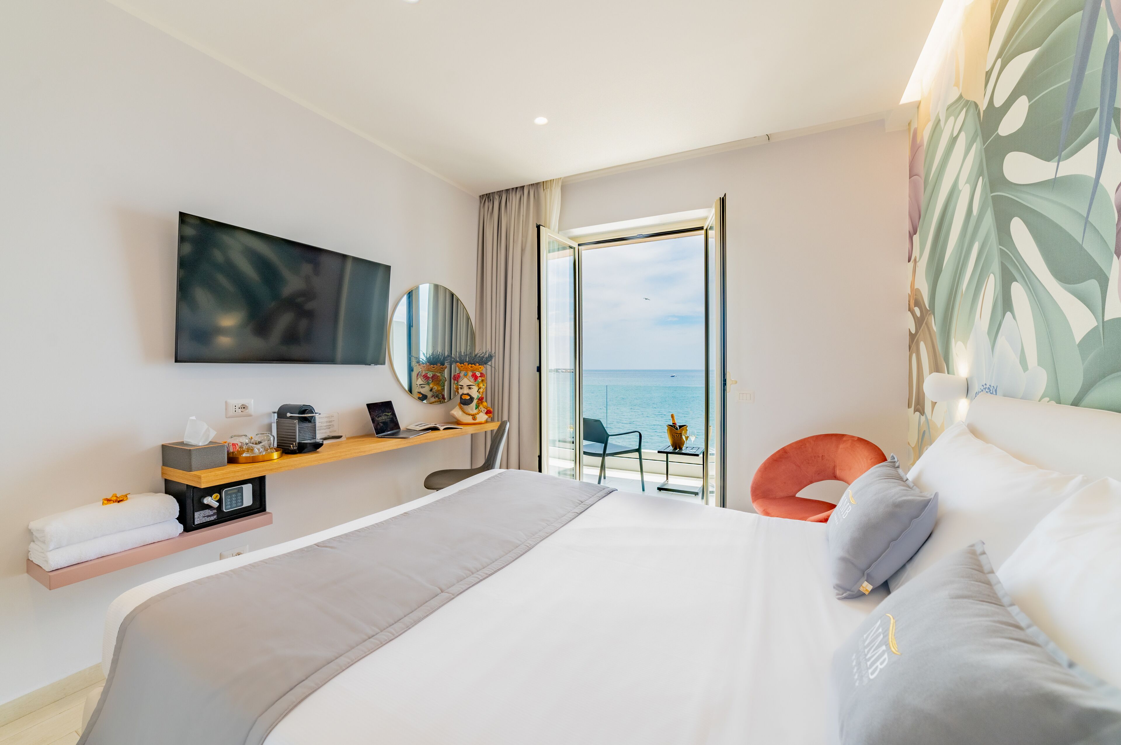 Quarto de hotel com uma cama grande, televisão na parede, secretária com computador e vista para o mar pelas portas da varanda.