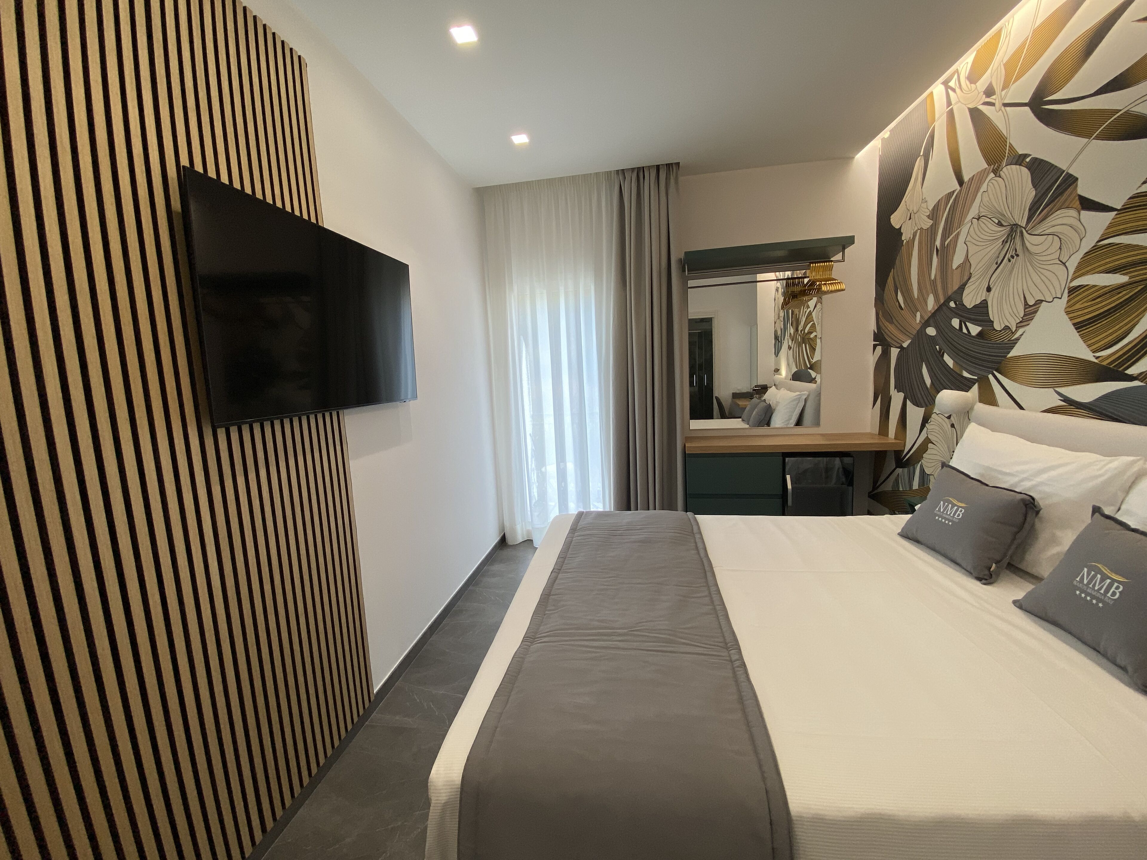 Una habitación de hotel moderna con una cama grande, un televisor de pantalla plana montado en la pared, un espejo y una pared decorativa con un estampado floral.