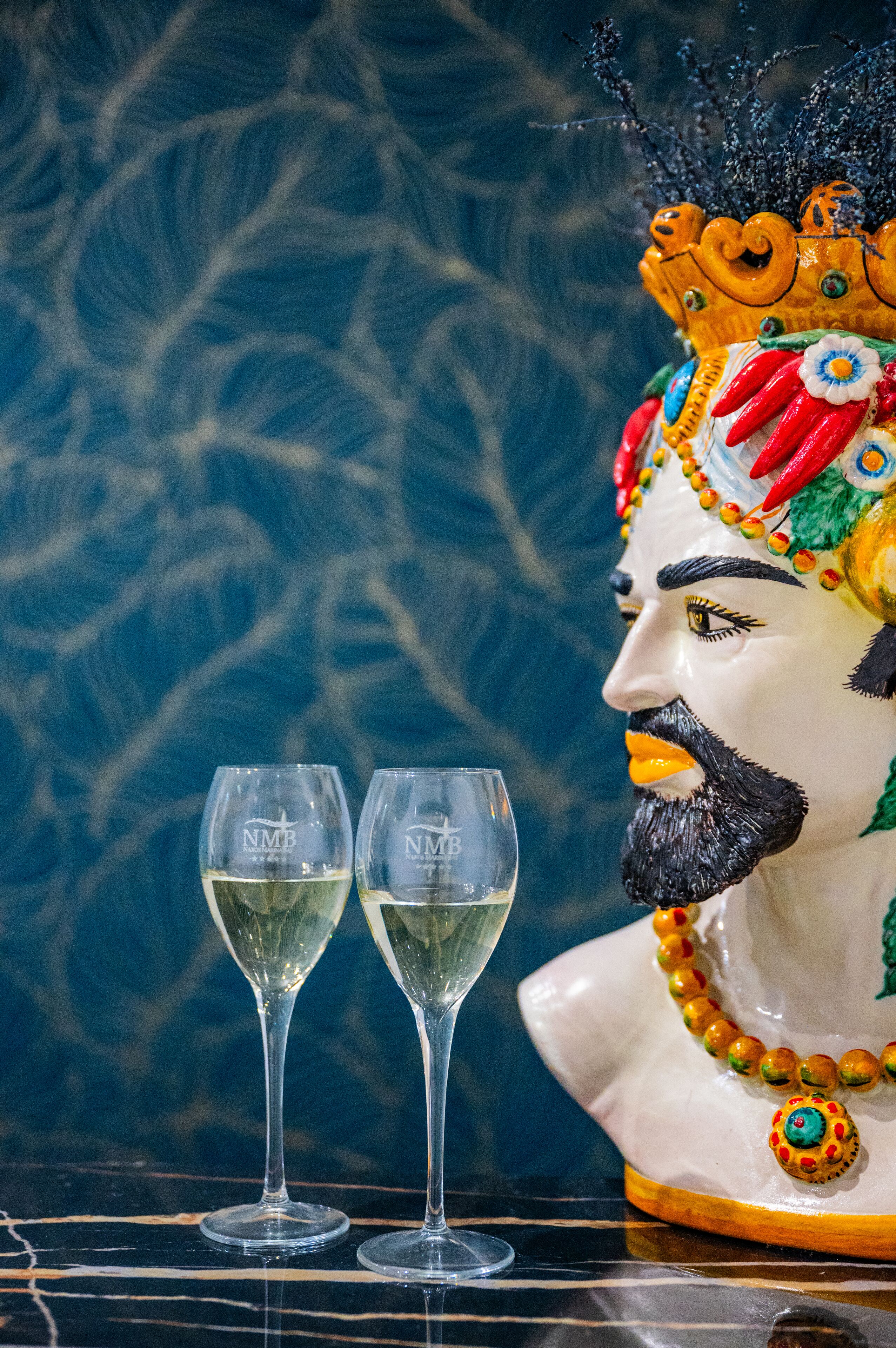 A statue with a colorful headdress and a white base, accompanied by two glasses of white wine on a reflective surface.