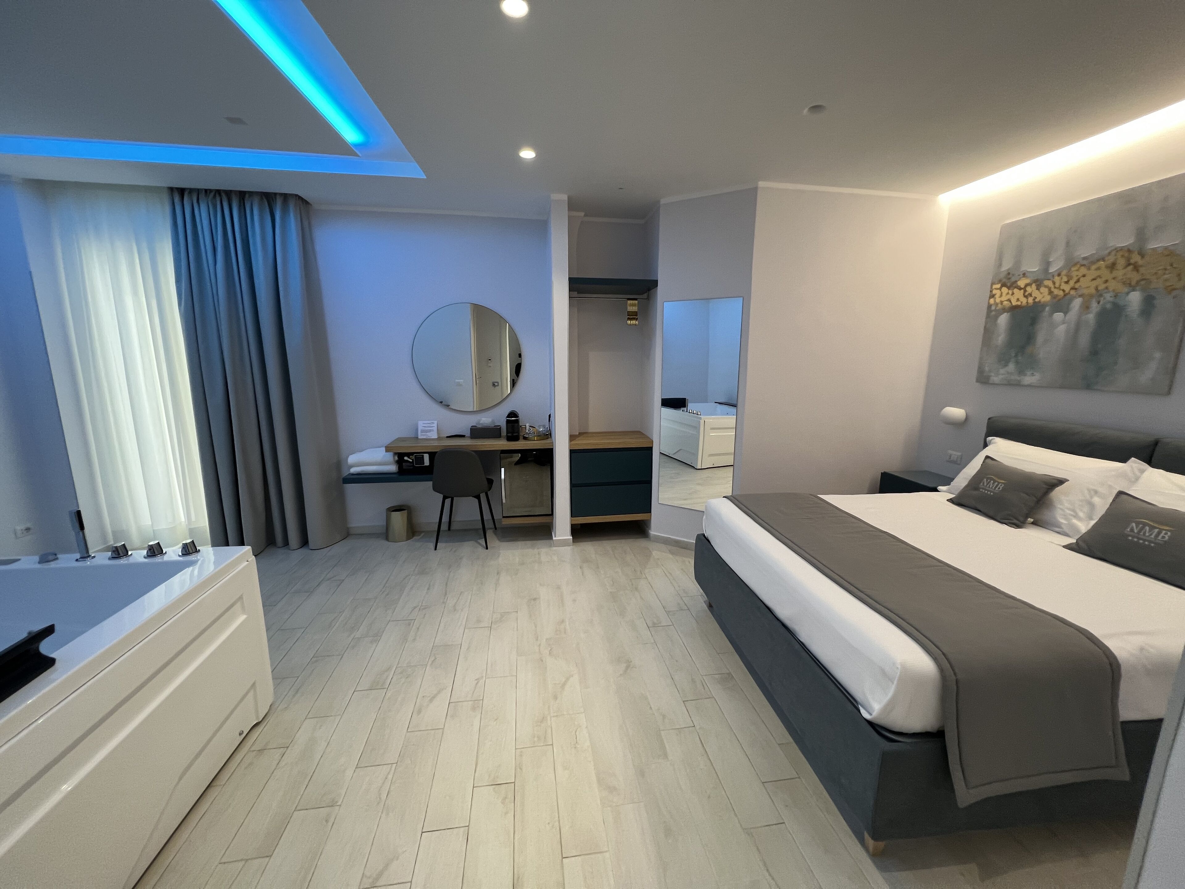 A modern hotel room with a large bed, a desk with a chair, a round mirror, and a bathroom with a bathtub.