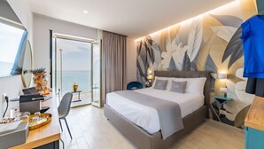 Une chambre d’hôtel moderne avec un grand lit, un bureau avec un ordinateur portable, une chaise et une vue sur la mer par une porte vitrée.